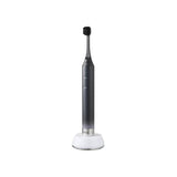 A sleek electric toothbrush with a dark gray handle and black bristles, resting on a white charging base against a solid white background. | Oral Care | YAMAN International